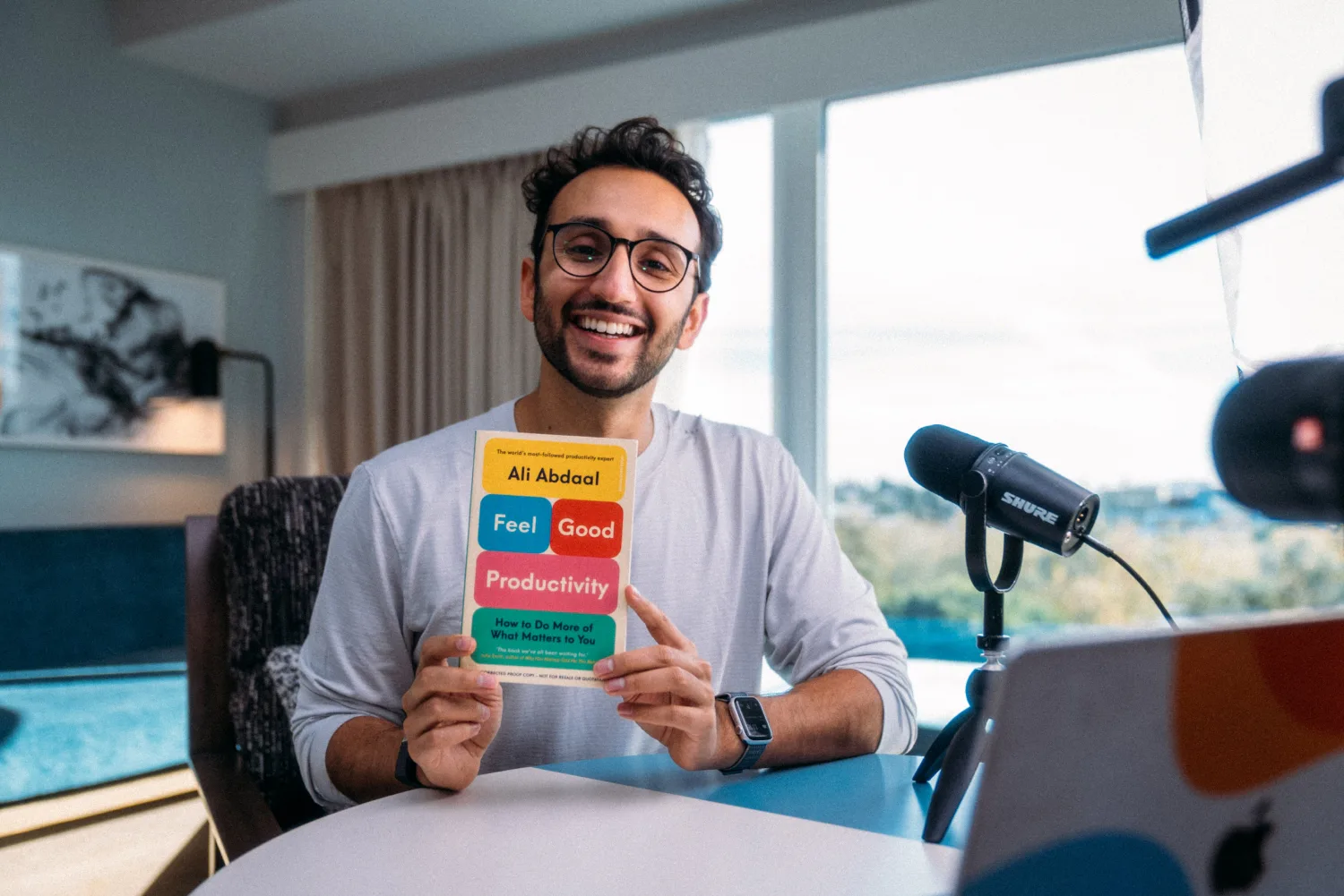 image of Ali Abdaal holding his book while sitting in front of a microphone and computer